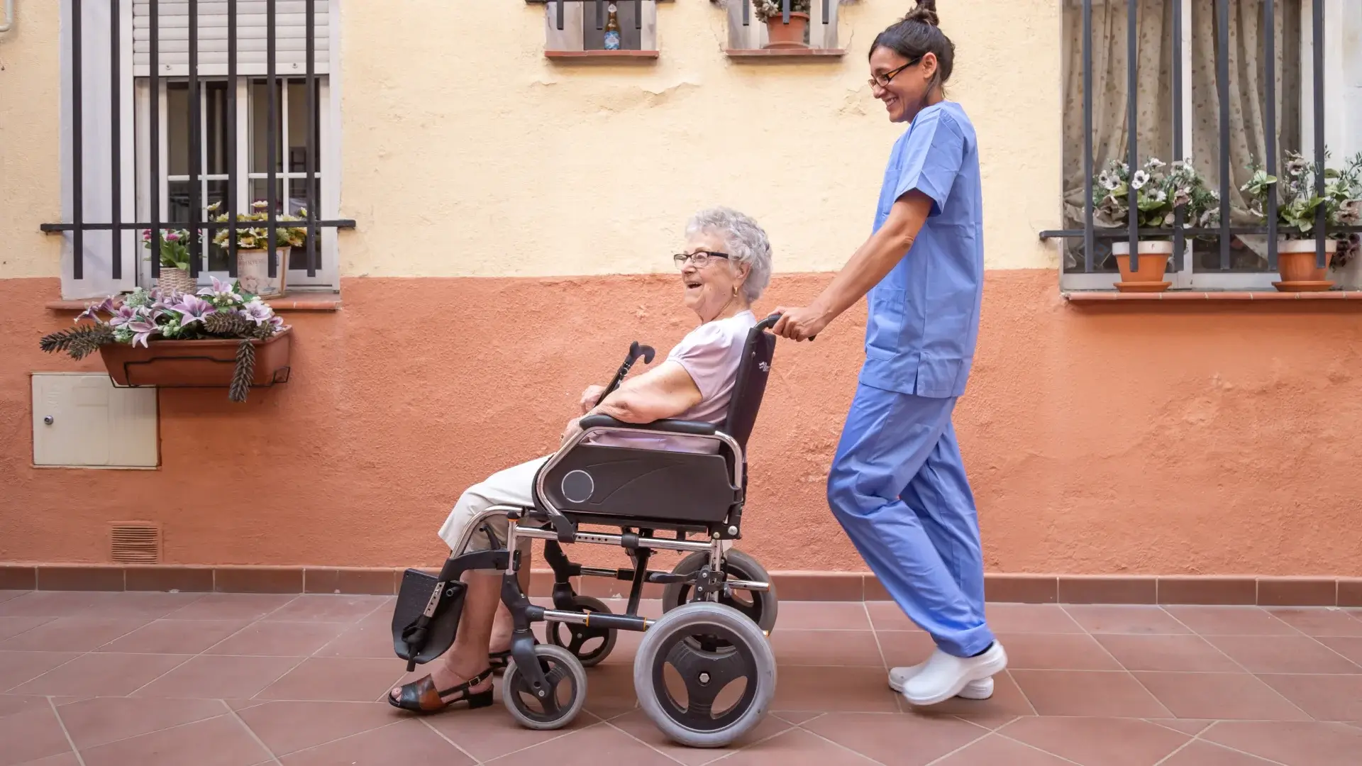 Caregiver pushes smiling elderly woman in wheelchair outdoors
